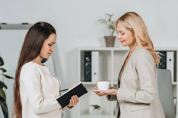 Side view of smiling business mentor with cup of coffee looking at young female colleague taking notes in notebook — Stock Photo