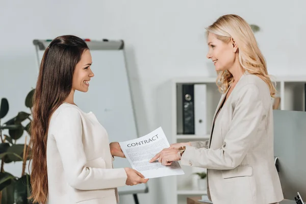 Боковой вид жизнерадостных деловых женщин, держащих контракт и улыбающихся друг другу в офисе — стоковое фото