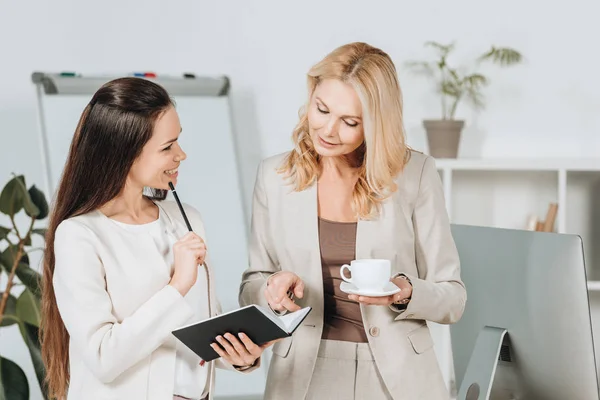 Улыбающиеся деловые женщины с чашкой кофе и блокнотом, стоящие вместе и обсуждающие работу в офисе — стоковое фото