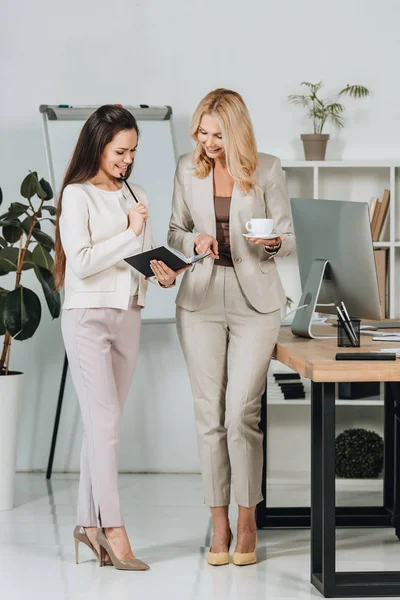 Belles femmes d'affaires heureuses avec tasse de café et cahier debout ensemble et discuter du travail au bureau — Photo de stock