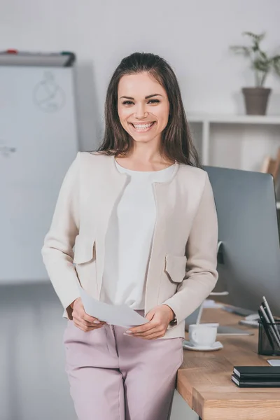 Bela jovem empresária feliz segurando papéis e sorrindo para a câmera no escritório — Fotografia de Stock