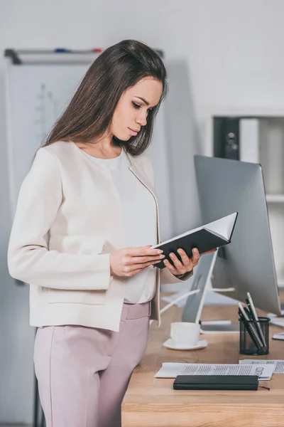 Konzentrierte junge Geschäftsfrau steht und hält Notizbuch im Büro — Stockfoto