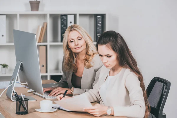 Mulher de negócios madura usando computador desktop e olhando para jovem colega trabalhando com papéis — Fotografia de Stock