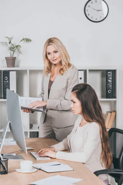 Mentor d'affaires mature avec dossier et jeune femme d'affaires regardant l'ordinateur de bureau dans le bureau — Photo de stock