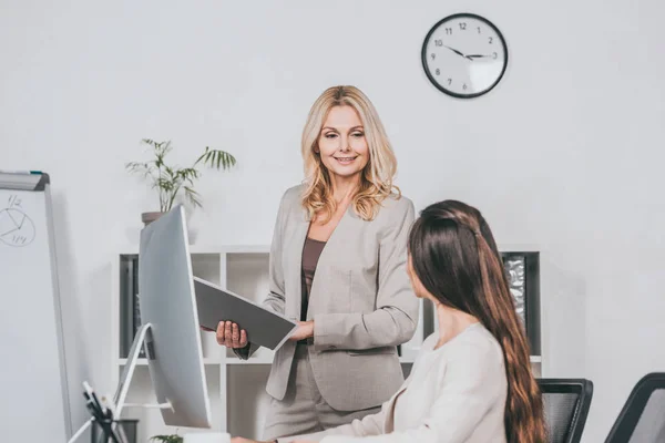 Sorridente donna d'affari matura che tiene la cartella e guarda il giovane collega che lavora con il computer desktop in ufficio — Foto stock