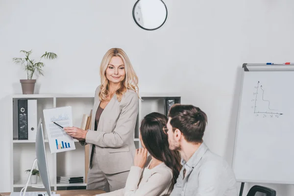 Smiling business mentor pointing at charts and looking at young colleagues sitting in office — Stock Photo