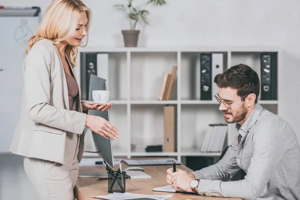 Lächelnde Geschäftsfrau, die eine Tasse Kaffee in der Hand hält und jungen Geschäftsmann mit Brille am Arbeitsplatz ansieht — Stockfoto