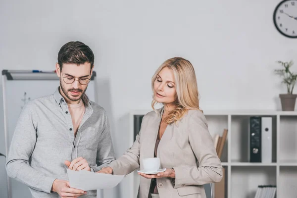 Жіночий бізнес-психолог тримає чашку кави і дивиться на молодого бізнесмена, який тримає документи в офісі — стокове фото