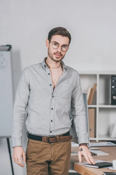 Jovem empresário profissional bonito em óculos de pé e olhando para a câmera no escritório — Fotografia de Stock