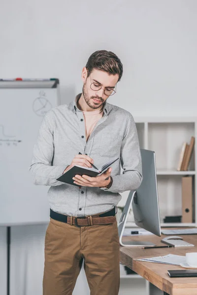 Fokussierter junger Geschäftsmann mit Brille im Büro stehend und schreibend im Notizbuch — Stockfoto