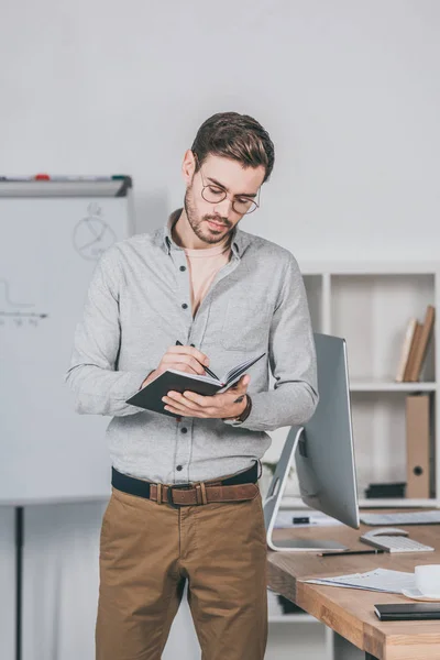 Focused young businessman in eyeglasses standing and writing in notebook in office — Stock Photo