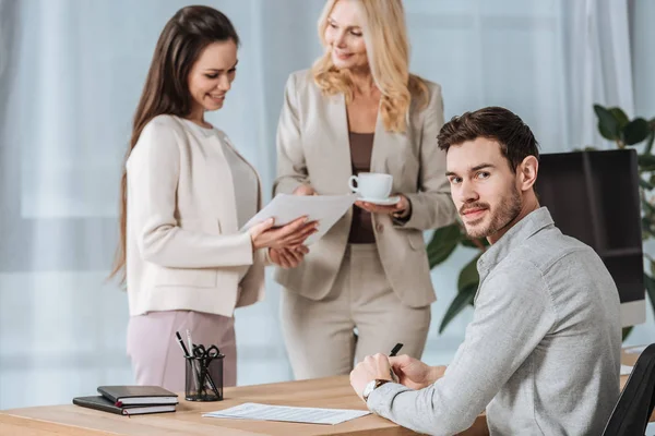 Donne d'affari sorridenti parlando e giovane uomo d'affari guardando la fotocamera in ufficio — Foto stock