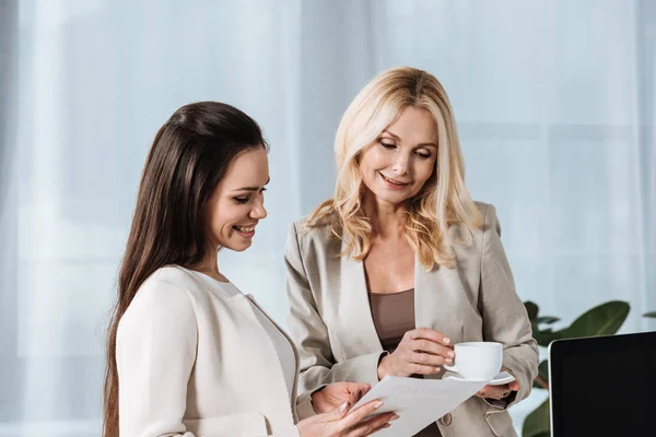 Lächelnde reife Geschäftsfrau, die eine Tasse Kaffee hält und junge Kollegin mit Papieren im Büro ansieht — Stockfoto
