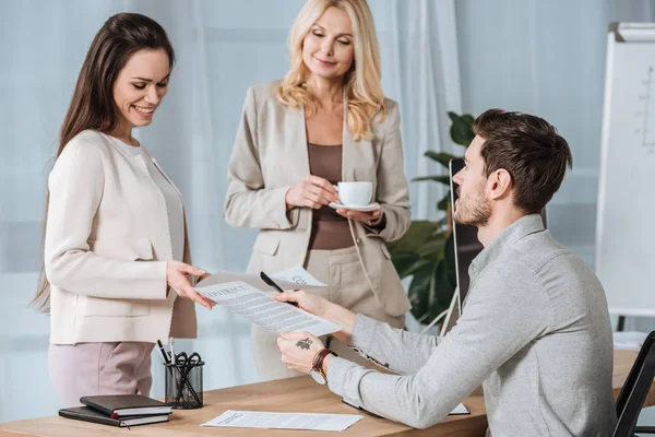 Lächelnde reife Geschäftsfrau mit einer Tasse Kaffee und Blick auf junge Kollegen, die im Büro mit Papieren arbeiten — Stockfoto