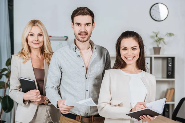 Professionelle reife Business-Mentorin mit jungen Kollegen, die im Büro zusammenstehen und in die Kamera lächeln — Stockfoto