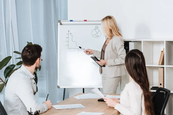 Mentor d'affaires pointant vers le tableau blanc avec des tableaux tandis que de jeunes collègues assis à la table dans le bureau — Photo de stock