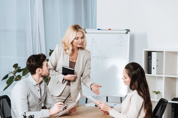 Femme d'affaires mature souriante regardant de jeunes collègues assis avec des papiers au bureau — Photo de stock