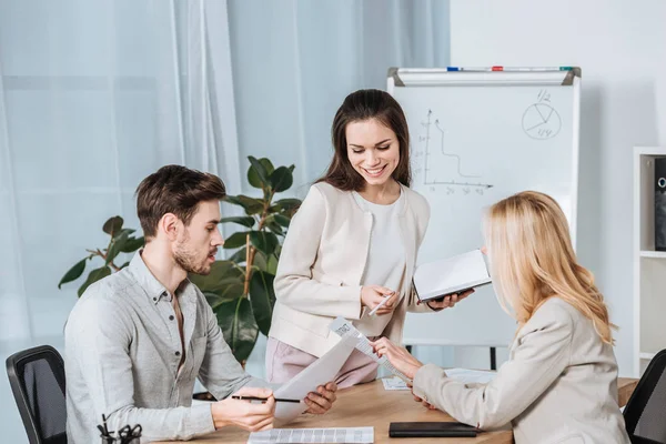 Lächelnde junge Geschäftsfrau mit Blick auf männliche und weibliche Kollegen, die mit Papieren am Schreibtisch im Büro sitzen — Stockfoto