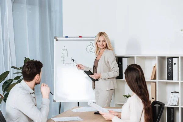 Lächelnde reife Geschäftsfrau zeigt auf Whiteboard und schaut junge Kollegen im Büro an — Stockfoto