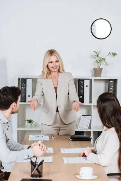 Sonriente mujer mentora de negocios mirando a jóvenes colegas sentados en el lugar de trabajo - foto de stock