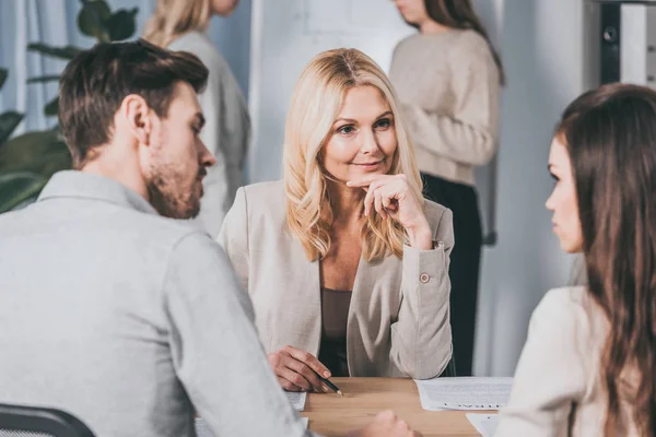 Schöner lächelnder Business-Mentor, der mit der Hand am Kinn sitzt und junge Kollegen im Büro betrachtet — Stockfoto