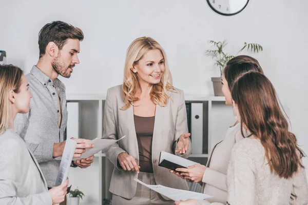 Jóvenes empresarios sosteniendo papeles y mirando al mentor sonriente profesional en el cargo - foto de stock