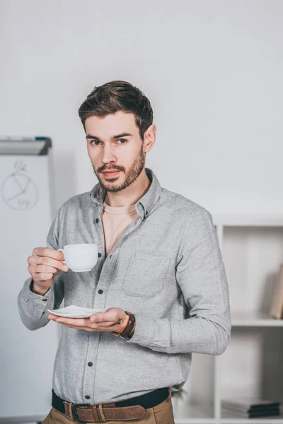 Handsome young businessman holding cup of coffee and looking at camera in office — Stock Photo