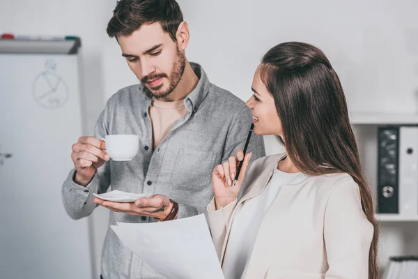 Business mentor holding cup of coffee and discussing papers with female colleague in office — Stock Photo