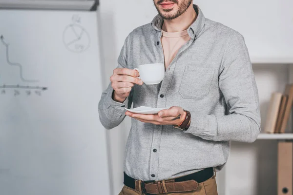 Tiro cortado de barbudo jovem empresário segurando xícara de café e pires no escritório — Fotografia de Stock