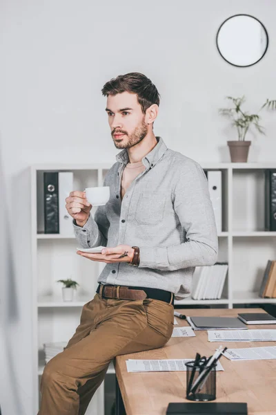 Young businessman holding cup of coffee and sitting on desk in office — Stock Photo
