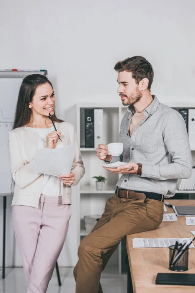 Lächelnde junge Geschäftsfrau hält Papiere in der Hand und unterhält sich mit hübschen männlichen Kollegen, die im Büro Kaffee trinken — Stockfoto