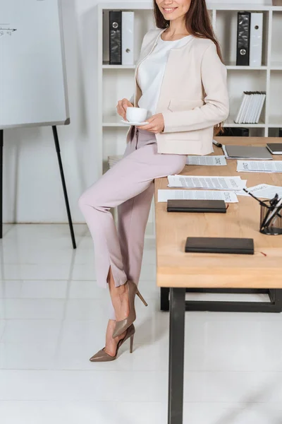 Schnappschuss einer lächelnden jungen Geschäftsfrau, die eine Tasse Kaffee hält und im Büro auf einem Tisch sitzt — Stockfoto