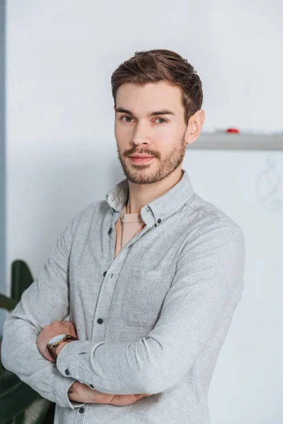 Confident young businessman standing with crossed arms and looking at camera in office — Stock Photo