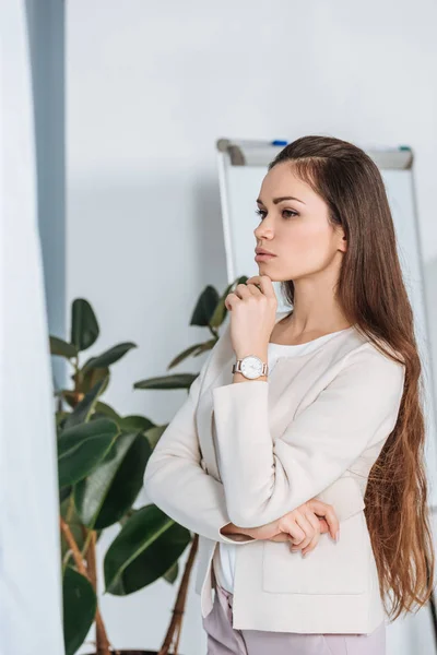 Réfléchie jeune femme d'affaires debout avec la main sur le menton et regardant loin dans le bureau — Photo de stock