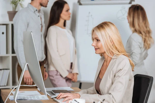 Vista lateral de mulher de negócios sorrindo usando computador desktop e jovens colegas que trabalham com quadro branco atrás — Fotografia de Stock