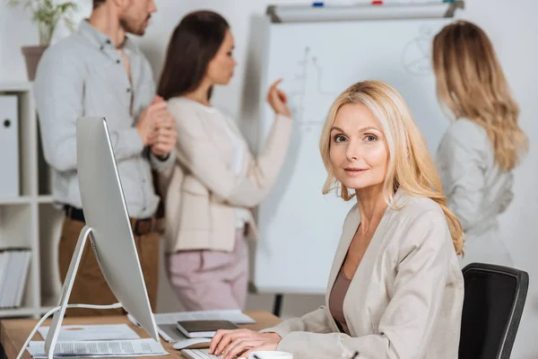 Mature femme d'affaires en utilisant un ordinateur de bureau et en regardant la caméra tandis que les jeunes collègues travaillent avec tableau blanc derrière — Photo de stock