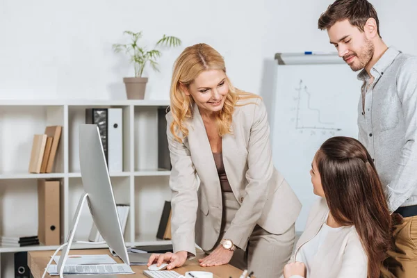 Smiling mature businesswoman using desktop computer and looking at young colleagues in office — Stock Photo