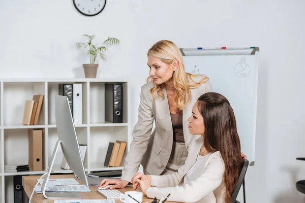 Giovane donna d'affari seduta alla scrivania e guardando sorridente mentore femminile utilizzando il computer desktop in ufficio — Foto stock