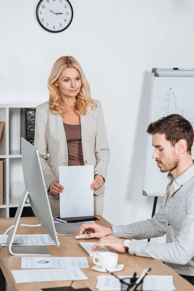 Mentor de negocios maduro sosteniendo papeles y mirando al joven hombre de negocios utilizando la computadora de escritorio en la oficina — Stock Photo