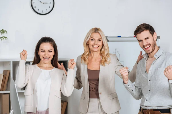 Веселые профессиональные деловые коллеги, трясущие кулаками и улыбающиеся перед камерой в офисе — стоковое фото