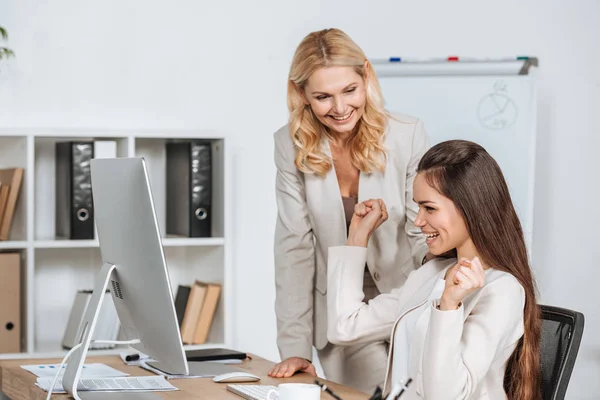 Souriant femme d'affaires mature regardant jeune collègue joyeux en utilisant un ordinateur de bureau sur le lieu de travail — Photo de stock