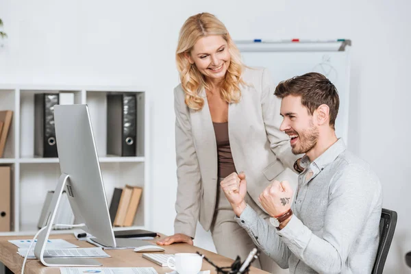 Sonriente mentor femenina mirando feliz joven hombre de negocios trabajando con computadora de escritorio en la oficina - foto de stock