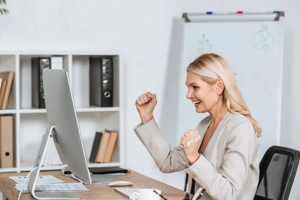 Cheerful mature businesswoman triumphing and using desktop computer at workplace — Stock Photo