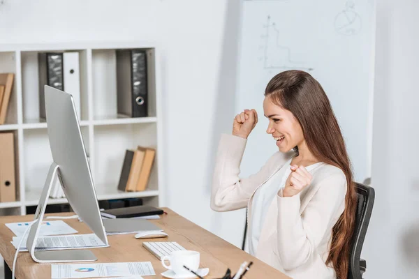 Seitenansicht einer glücklichen jungen Geschäftsfrau, die im Büro die Fäuste schüttelt und auf den Desktop-Computer blickt — Stockfoto