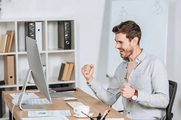 Aufgeregter junger Geschäftsmann schüttelt im Büro die Fäuste und blickt auf den Desktop-Computer — Stockfoto