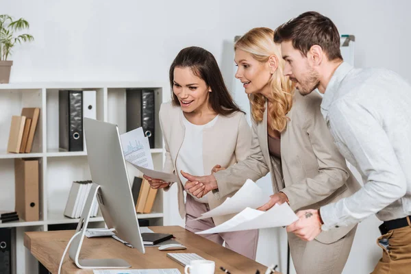 Mentor de negocios sonriente con colegas jóvenes sosteniendo papeles y usando computadora de escritorio en la oficina - foto de stock