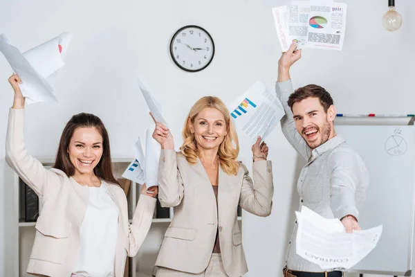 Веселые профессиональные деловые коллеги, держащие бумаги и улыбающиеся перед камерой в офисе — стоковое фото
