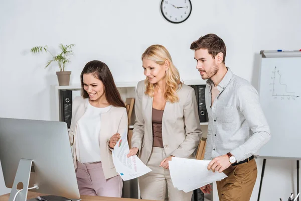 Sonrientes empresarios profesionales sosteniendo papeles y mirando a la computadora de escritorio en la oficina - foto de stock