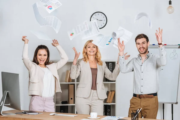 Empresarios profesionales emocionados lanzando papeles y sonriendo a la cámara en la oficina - foto de stock