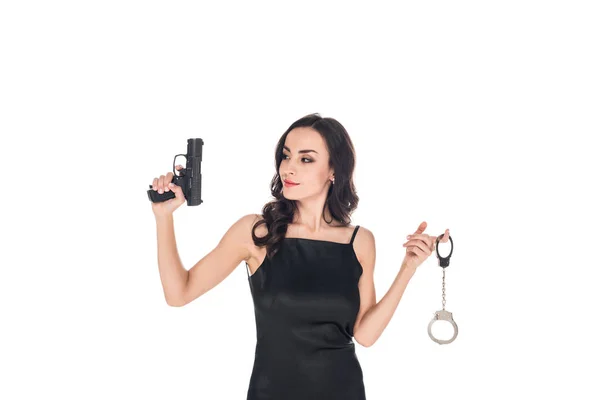 Atraente agente secreto elegante em vestido preto segurando arma e algemas, isolado em branco — Fotografia de Stock